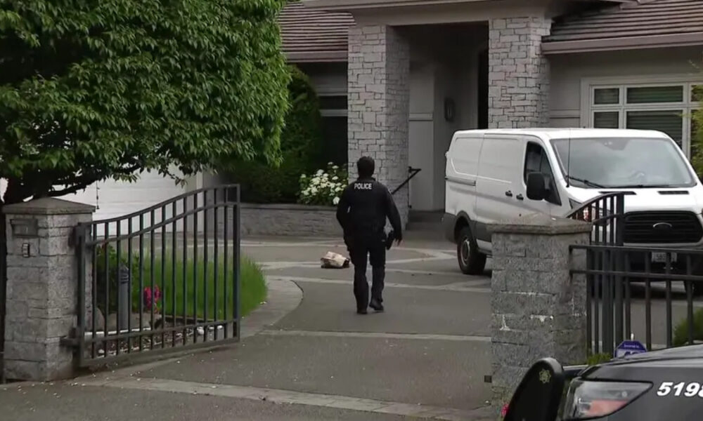 Police officer in a dark uniform walks toward a residential driveway with a white cargo van parked near a stone house, gate in foreground.
