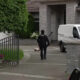 Police officer in a dark uniform walks toward a residential driveway with a white cargo van parked near a stone house, gate in foreground.
