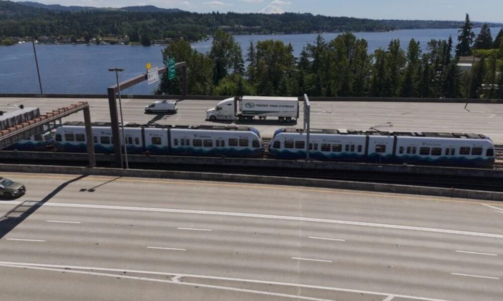 Passenger train on elevated tracks above a multi-lane highway, with a Frozen Food Express truck on the platform and a lake and trees in the background.