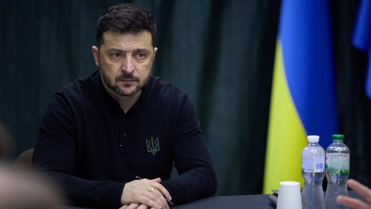 Man with short dark hair and beard sits at a table, wearing a black shirt with a Ukrainian emblem; Ukrainian flag in the background.