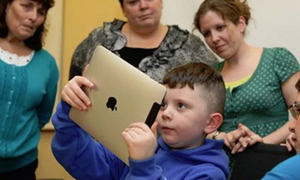 Boy in a blue hoodie holds a gold tablet up, surrounded by adults in a classroom setting, watching him.