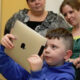 Boy in a blue hoodie holds a gold tablet up, surrounded by adults in a classroom setting, watching him.