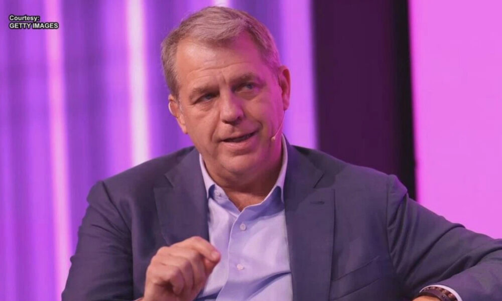Man in a blue blazer and light blue shirt sits on a purple-lit stage, speaking with a headset mic on a panel backdrop.