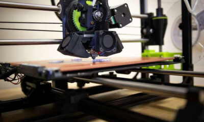 A 3D printer extruding material onto a pinkish print bed with a black printhead and gears in view, in a workshop setting.