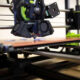 A 3D printer extruding material onto a pinkish print bed with a black printhead and gears in view, in a workshop setting.