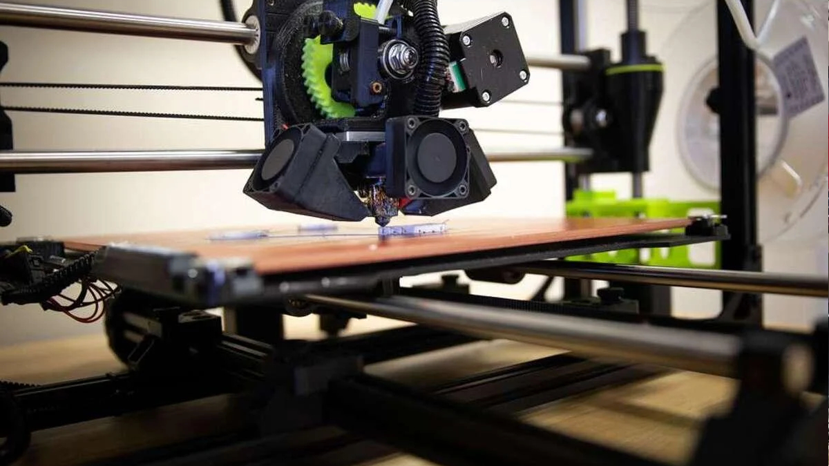 A 3D printer extruding material onto a pinkish print bed with a black printhead and gears in view, in a workshop setting.