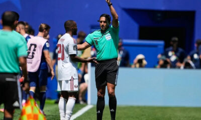 Soccer referee in teal shirt raises an arm while speaking to a player in a white jersey near the sideline.