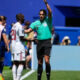 Soccer referee in teal shirt raises an arm while speaking to a player in a white jersey near the sideline.