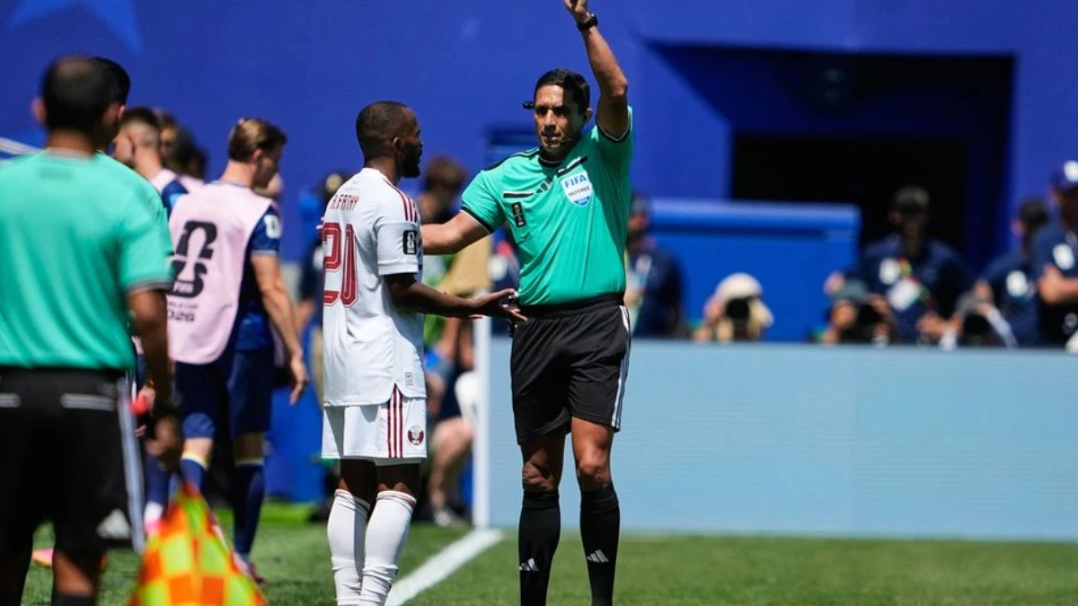 Soccer referee in teal shirt raises an arm while speaking to a player in a white jersey near the sideline.