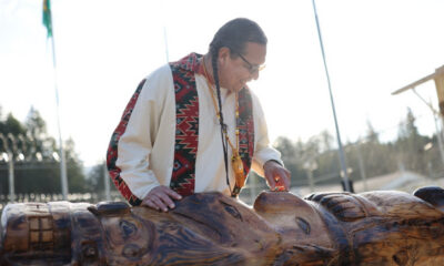 Indigenous craftsman with braids and beaded necklace carves a large wooden sculpture outdoors, sunlight behind him.