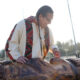 Indigenous craftsman with braids and beaded necklace carves a large wooden sculpture outdoors, sunlight behind him.