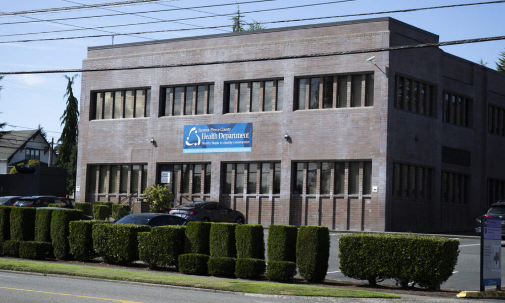 Brick two-story Health Department building with a blue sign reading Health Department, in a parking lot gated by trimmed hedges.