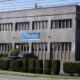 Brick two-story Health Department building with a blue sign reading Health Department, in a parking lot gated by trimmed hedges.