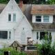 White two-story house with a steep front gable; leaves cover parts of the roof and a small animal in the front yard near a toy playhouse.