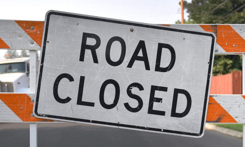 Road closed sign leaning on an orange-and-white construction barrier across a street, with TEXT: ROAD CLOSED clearly visible.