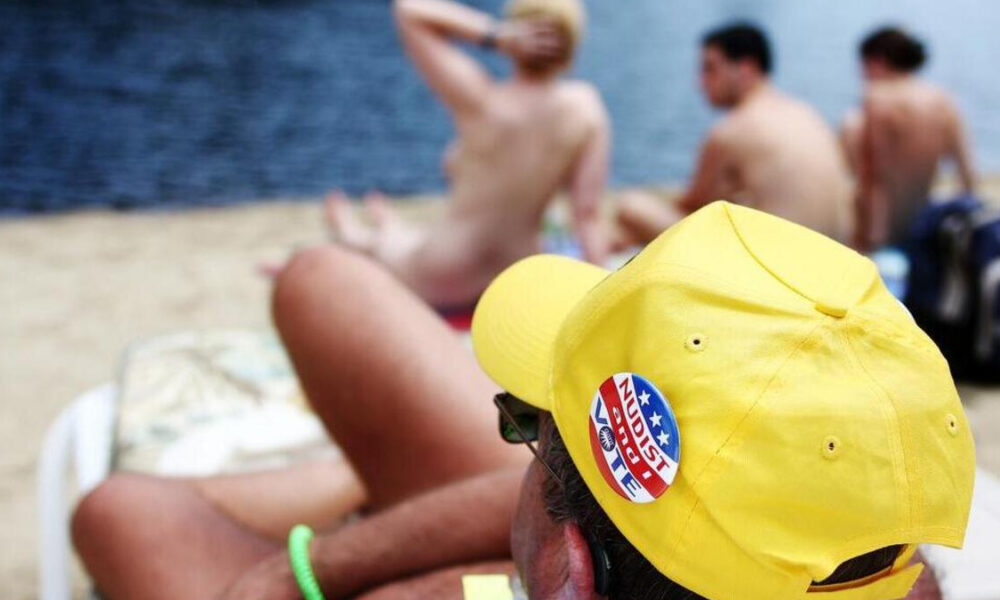 Back view of a man on a nude beach wearing a bright yellow cap with a Nudist badge, with topless sunbathers and the sea in the background.