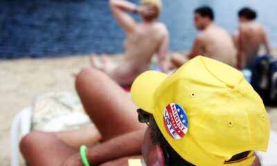 Back view of a man on a nude beach wearing a bright yellow cap with a Nudist badge, with topless sunbathers and the sea in the background.