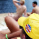 Back view of a man on a nude beach wearing a bright yellow cap with a Nudist badge, with topless sunbathers and the sea in the background.