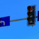 Traffic light signals at an intersection with a left-turn sign and a right/straight lane sign against a blue sky.