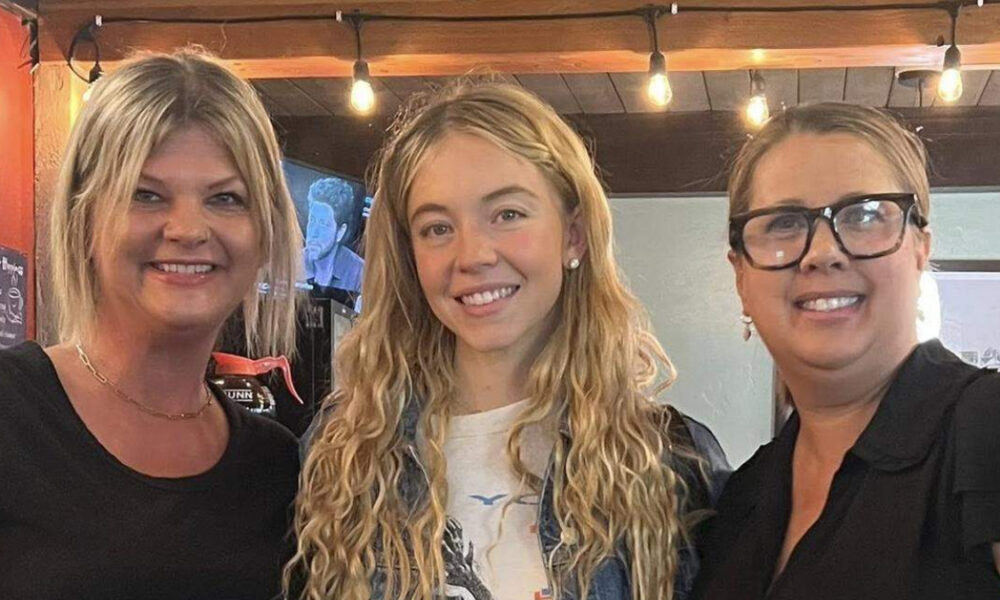 Three smiling women posing together indoors under warm string lights.