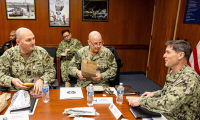 Three U.S. Navy sailors in camouflage sit at a conference table, one holding a brown paper bag.