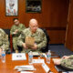Three U.S. Navy sailors in camouflage sit at a conference table, one holding a brown paper bag.