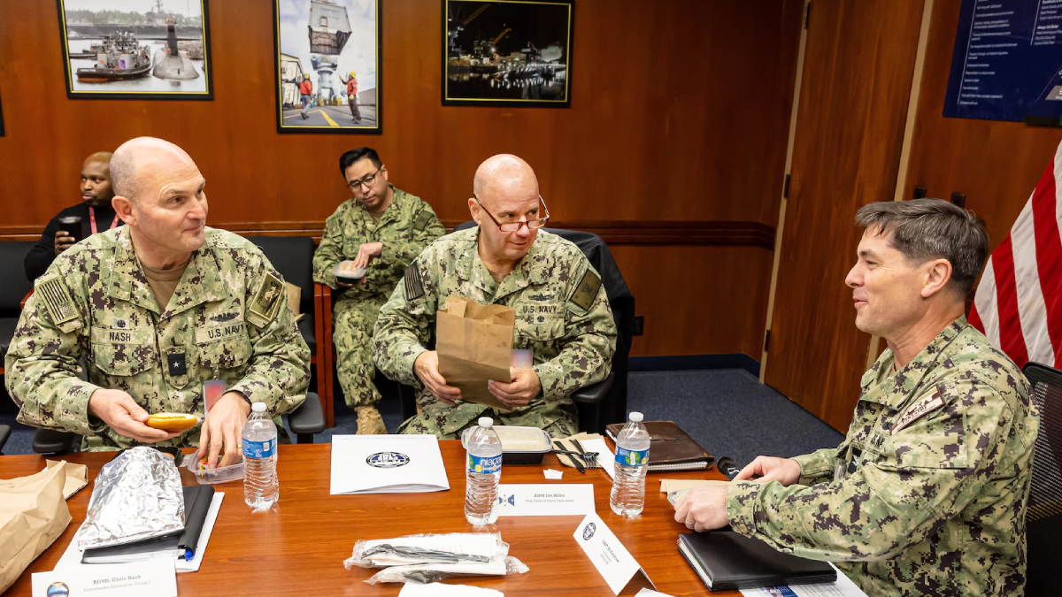Three U.S. Navy sailors in camouflage sit at a conference table, one holding a brown paper bag.