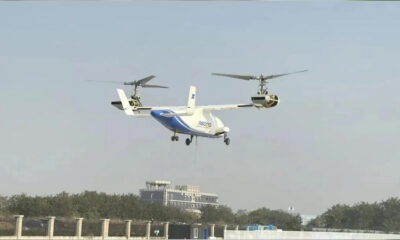 Twin-rotor VTOL aircraft hovering above a fence line near a building and trees.