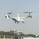 Twin-rotor VTOL aircraft hovering above a fence line near a building and trees.