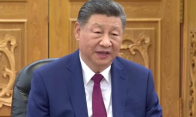 Man in a blue suit and red tie speaking in a formal room with ornate wooden panels behind him.