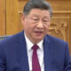 Man in a blue suit and red tie speaking in a formal room with ornate wooden panels behind him.