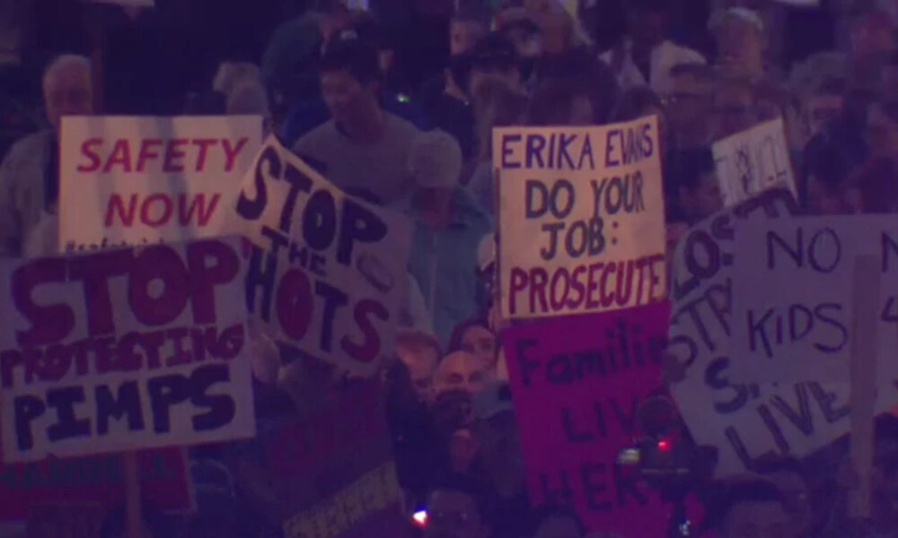Crowd of protesters at a rally holding handwritten signs, including 'SAFETY NOW' and 'STOP THE SHOTS'.