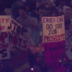 Crowd of protesters at a rally holding handwritten signs, including 'SAFETY NOW' and 'STOP THE SHOTS'.