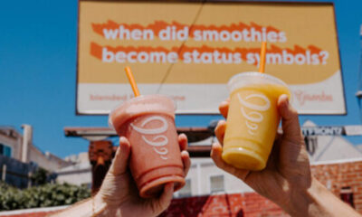 Two hands raise pink and yellow smoothies in front of a billboard that asks, 'when did smoothies become status symbols?''','Hands holding a pink smoothie and a yellow smoothie up to camera with a blue sky and an orange sign in the background reading about smoothies as status symbols.