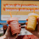 Two hands raise pink and yellow smoothies in front of a billboard that asks, 'when did smoothies become status symbols?''','Hands holding a pink smoothie and a yellow smoothie up to camera with a blue sky and an orange sign in the background reading about smoothies as status symbols.