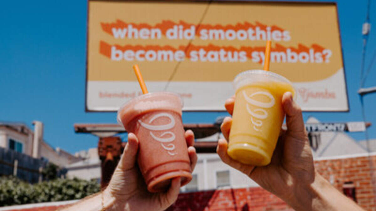 Two hands raise pink and yellow smoothies in front of a billboard that asks, 'when did smoothies become status symbols?''','Hands holding a pink smoothie and a yellow smoothie up to camera with a blue sky and an orange sign in the background reading about smoothies as status symbols.