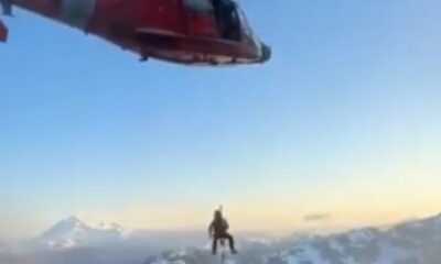 Red helicopter hovering above a lone climber on a snow-covered mountain ridge, clear blue sky in the background and distant peaks.