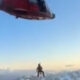 Red helicopter hovering above a lone climber on a snow-covered mountain ridge, clear blue sky in the background and distant peaks.