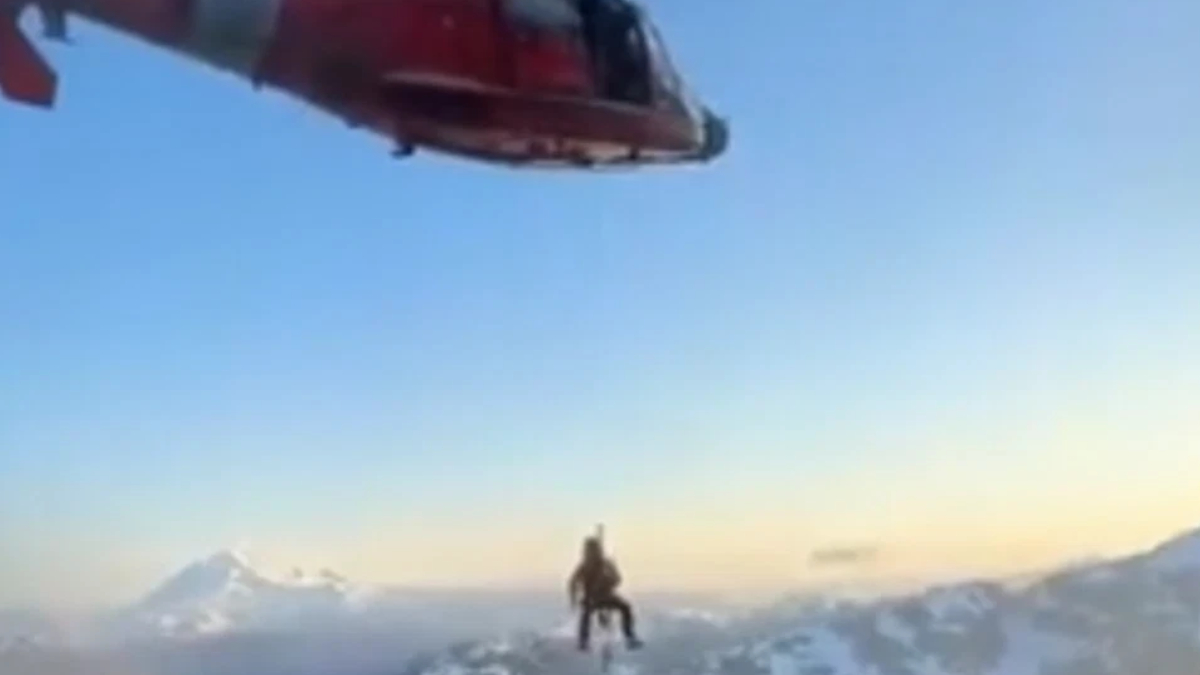 Red helicopter hovering above a lone climber on a snow-covered mountain ridge, clear blue sky in the background and distant peaks.