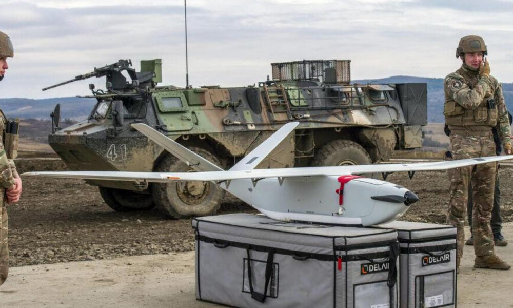 Two soldiers in camouflage stand beside a white fixed-wing drone on a gray equipment case, with a military vehicle in the background.