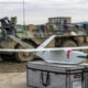 Two soldiers in camouflage stand beside a white fixed-wing drone on a gray equipment case, with a military vehicle in the background.