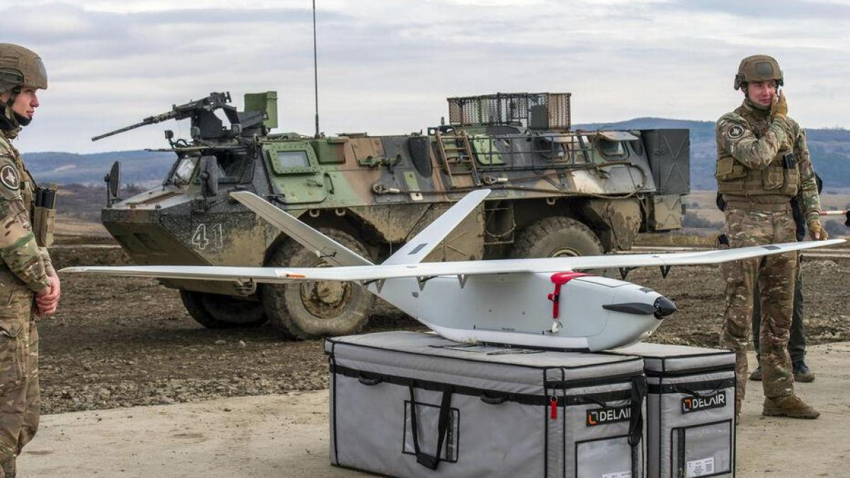 Two soldiers in camouflage stand beside a white fixed-wing drone on a gray equipment case, with a military vehicle in the background.