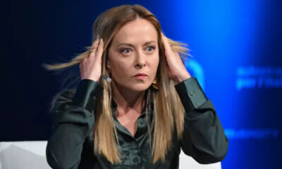 Woman on a panel with hands at her temples, a focused expression, and blue stage lighting in the background, wearing a dark blouse.