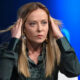 Woman on a panel with hands at her temples, a focused expression, and blue stage lighting in the background, wearing a dark blouse.