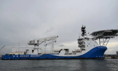 US Coast Guard cutter docked at a port with cranes and a large deck structure, blue hull and white superstructure.
