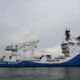 US Coast Guard cutter docked at a port with cranes and a large deck structure, blue hull and white superstructure.