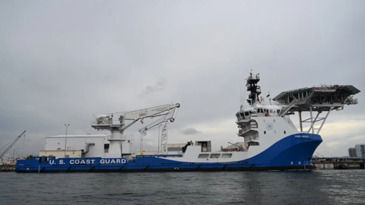 US Coast Guard cutter docked at a port with cranes and a large deck structure, blue hull and white superstructure.