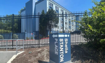Blue sign with white text and a left-pointing arrow reading '... Place' in front of a tall black fence and a light-blue apartment building behind it.