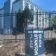 Blue sign with white text and a left-pointing arrow reading '... Place' in front of a tall black fence and a light-blue apartment building behind it.