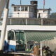 Harbor scene with a blue-and-white crane and a large white boat, workers in gear resting near a fence on the dock.